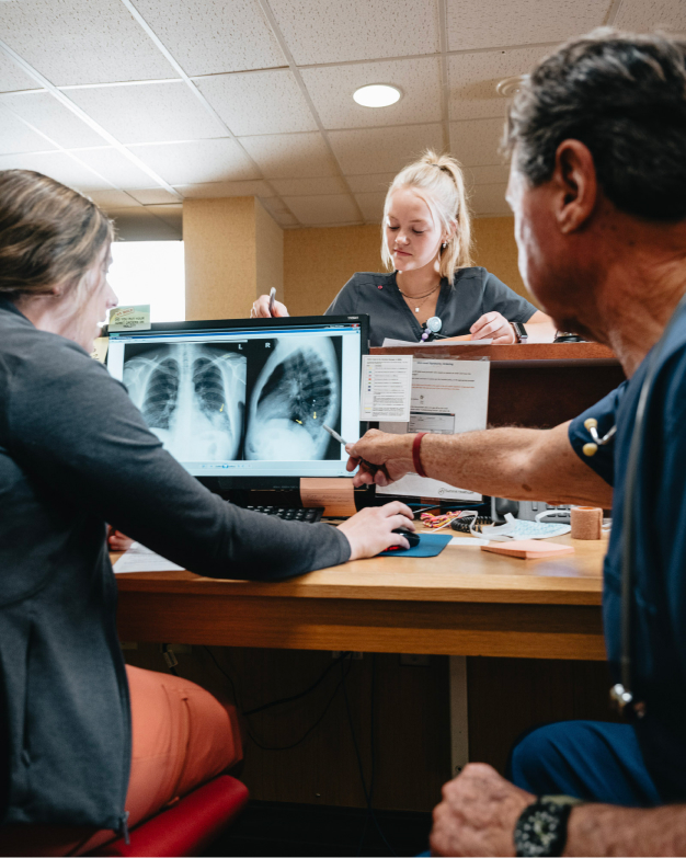 Three medical professionals discuss chest X-rays on a computer screen in a clinic setting.