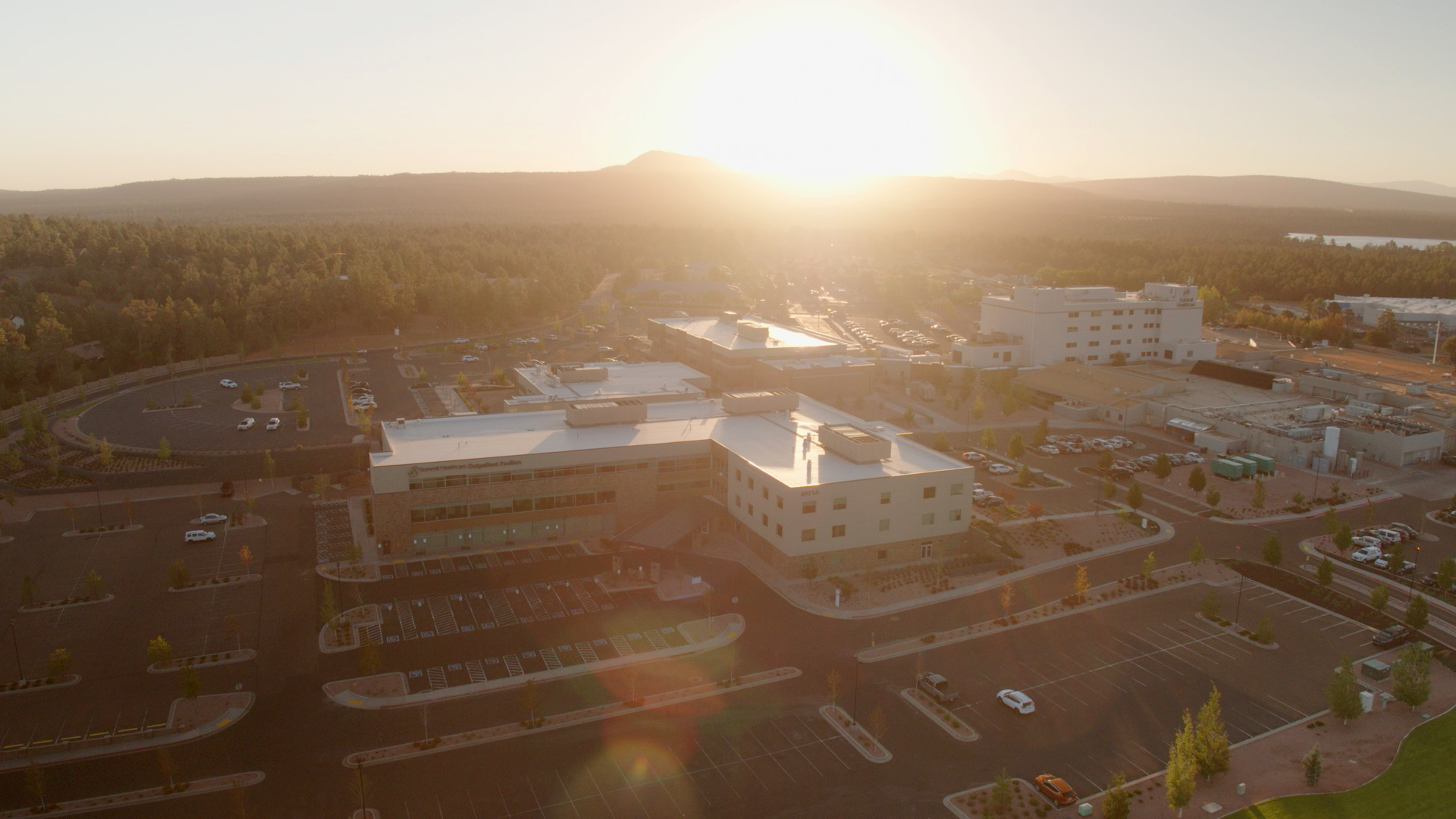 Aerial view of a hospital complex at sunset, surrounded by parking lots and trees, with a mountain range in the background.